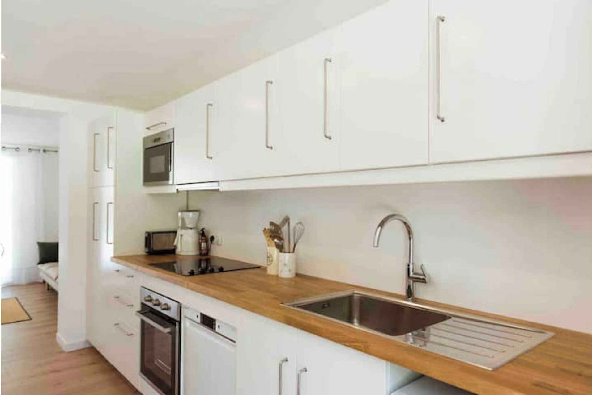 A modern kitchen with white cabinets, a wooden countertop, a built-in oven, a dishwasher, and a stainless steel sink.