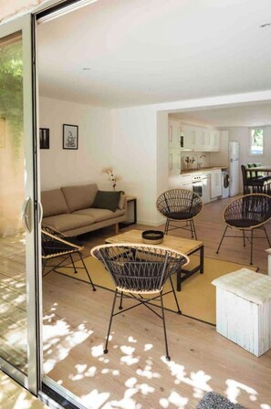 Un salon moderne comprenant un canapé, deux fauteuils en osier, une petite table et une porte en verre donnant sur un espace cuisine.