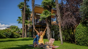 A couple on a blanket with a picnic setup, palm trees, and a modern building in the background. - Treelodgy (Riva del Garda)