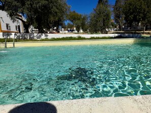 A clear swimming pool with a tiled edge, surrounded by trees and a white building with balconies. - Hacienda Marcharavi (Arcos de la Frontera)