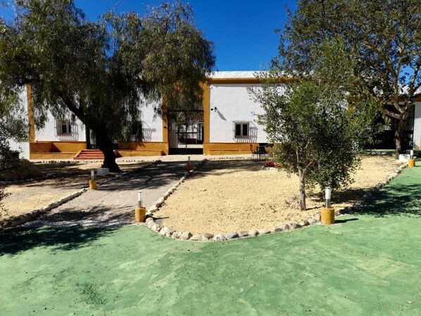 A building with a white facade and yellow trim, surrounded by trees and a gravel courtyard. - Hacienda Marcharavi (Arcos de la Frontera)