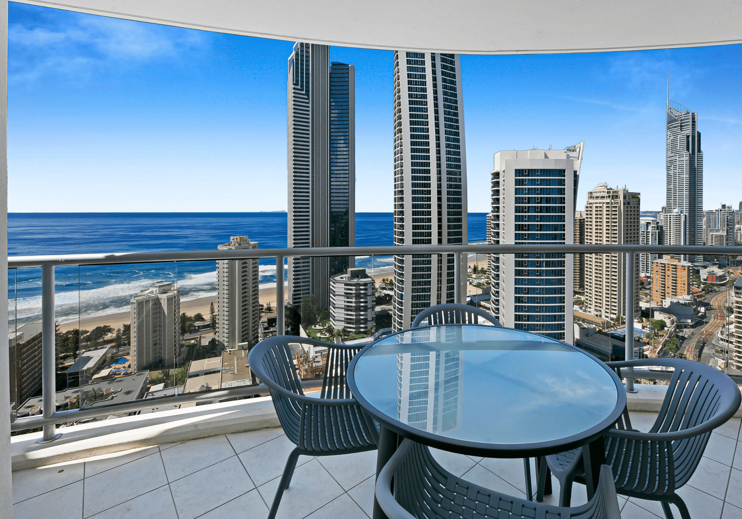Un balcon avec une table et des chaises en verre, offrant une vue sur le paysage urbain et la plage.