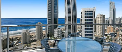 A balcony with a glass table and chairs, overlooking a cityscape and beach.
