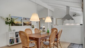 Un comedor con mesa de madera y sillas de mimbre, paredes blancas y techo con vigas a la vista.
