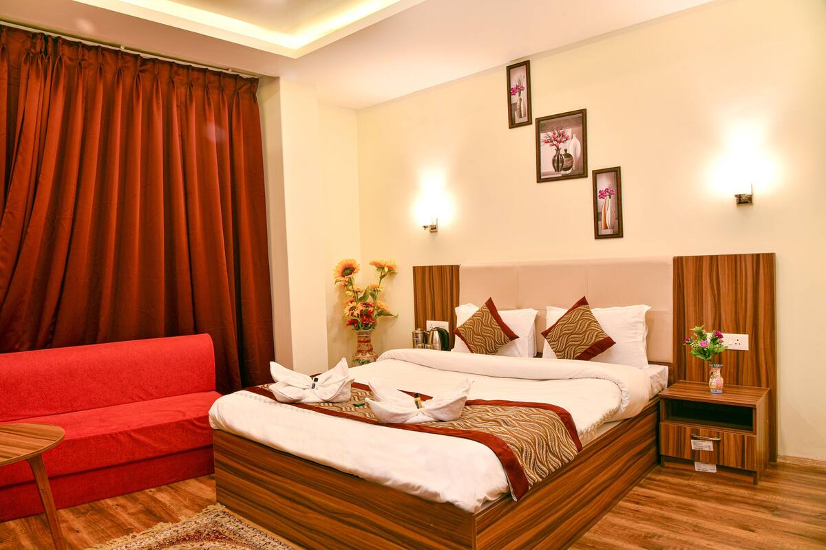 A hotel room with a large bed, a red sofa, a wooden nightstand, and a wall-mounted light fixture.