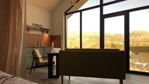 A bedroom with a large window, a bed, a chair, a small table, and a kitchen area with shelves and a sink.