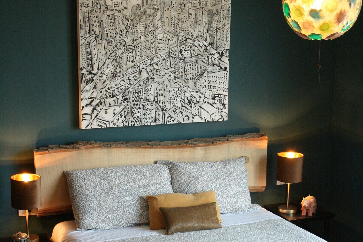 A neatly made bed with patterned bedding, a cityscape artwork above, and a hanging lamp with a colorful pattern.