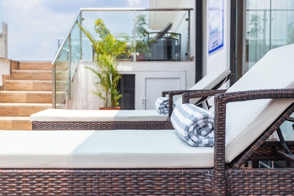 A wicker lounge chair with a white cushion and a striped towel, positioned near a staircase leading to a glass balcony. - Hotel Lo Mustang (Kathmandu)