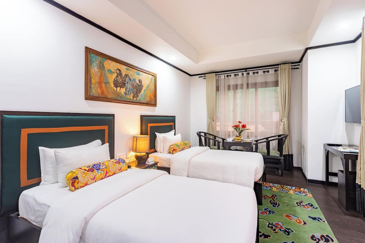 Habitación de hotel con dos camas, un escritorio, una silla, una mesa con un florero y un cuadro en la pared.