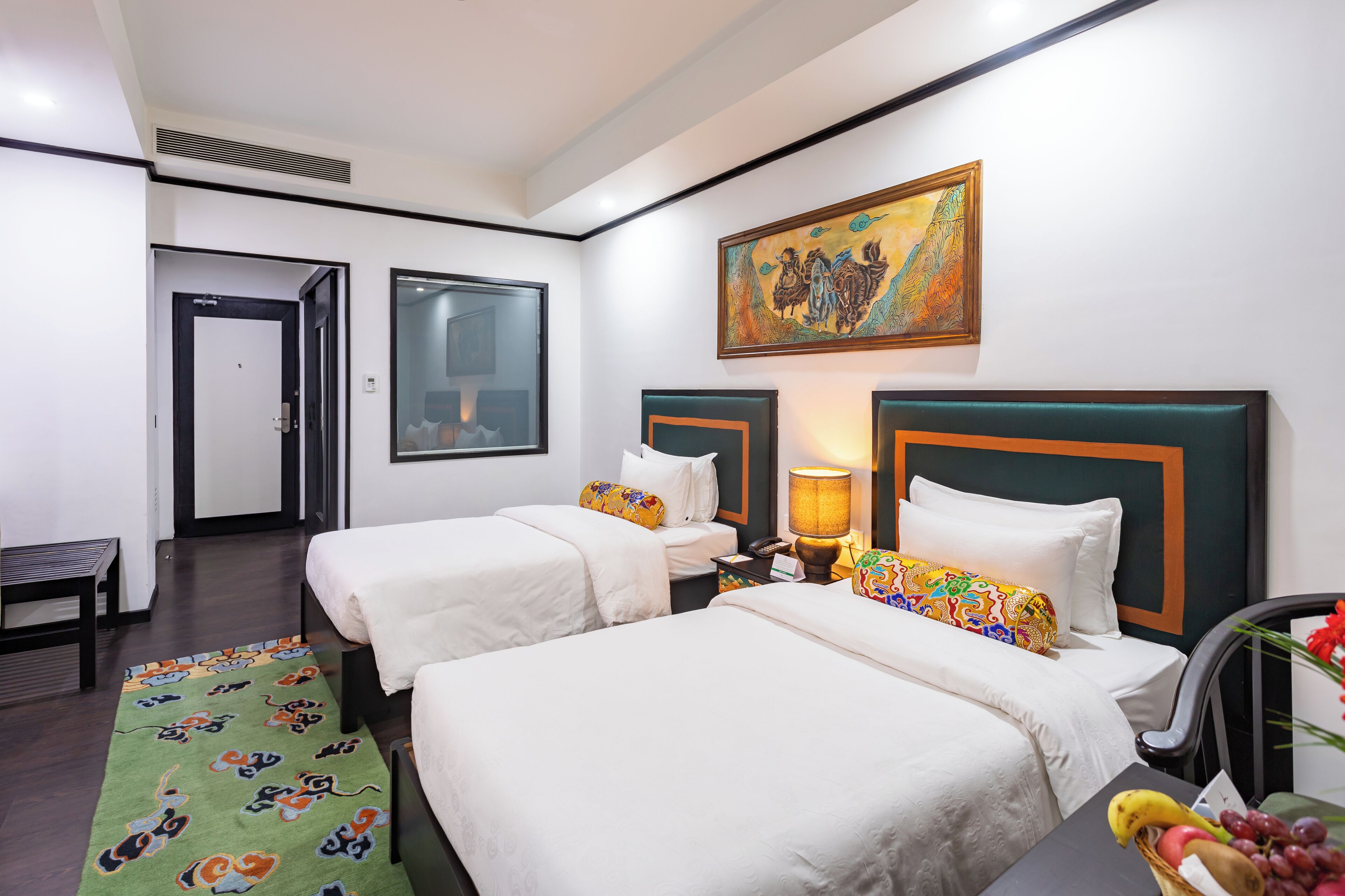 A hotel room with two single beds, a desk, a chair, a table with fruit, and a painting on the wall.