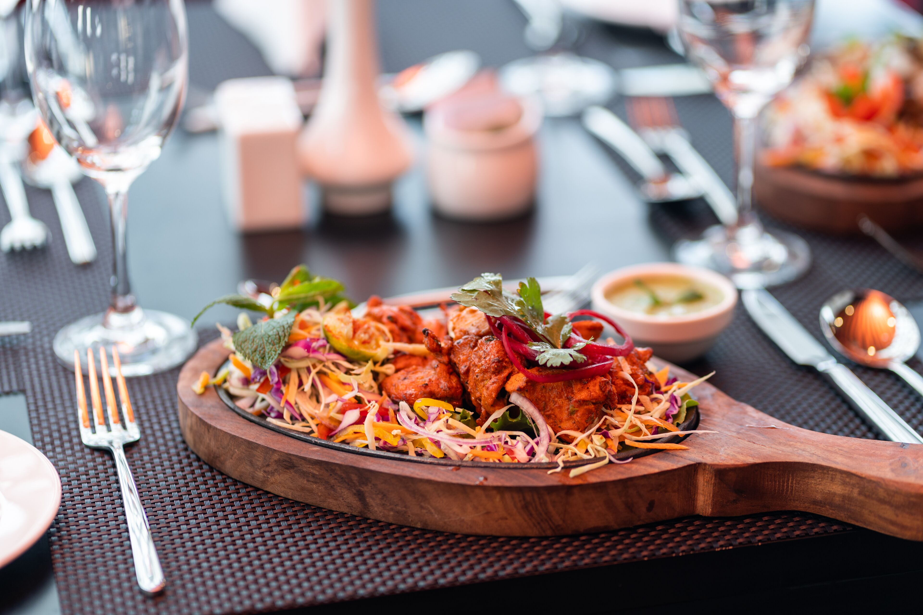 A wooden serving board with a colorful salad and meat dish, accompanied by cutlery and a glass of wine on a table.