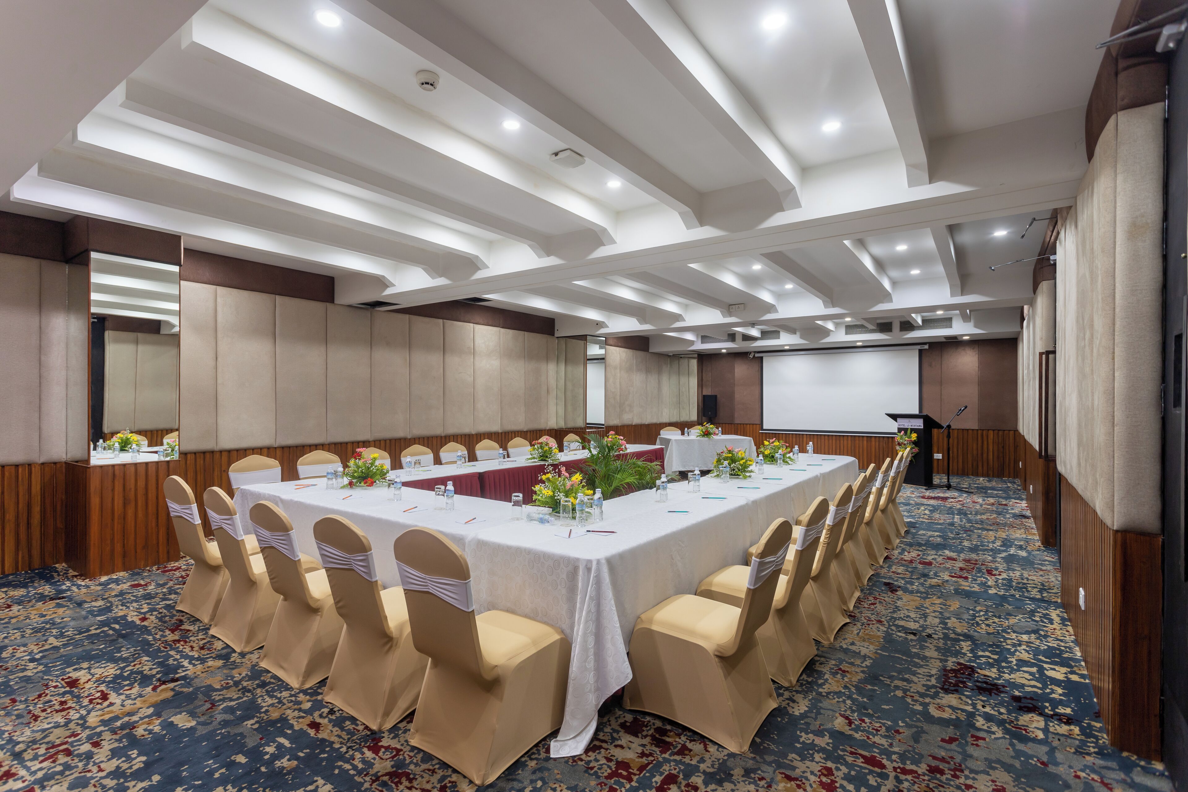 Un salón de conferencias con una larga mesa, sillas color beige y una pantalla para proyector.