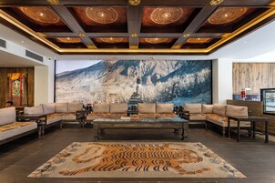 Um hall espaçoso com um grande mural na parede retratando uma paisagem montanhosa, uma área de estar central com sofás e uma mesa de centro, e um tapete estampado no chão.