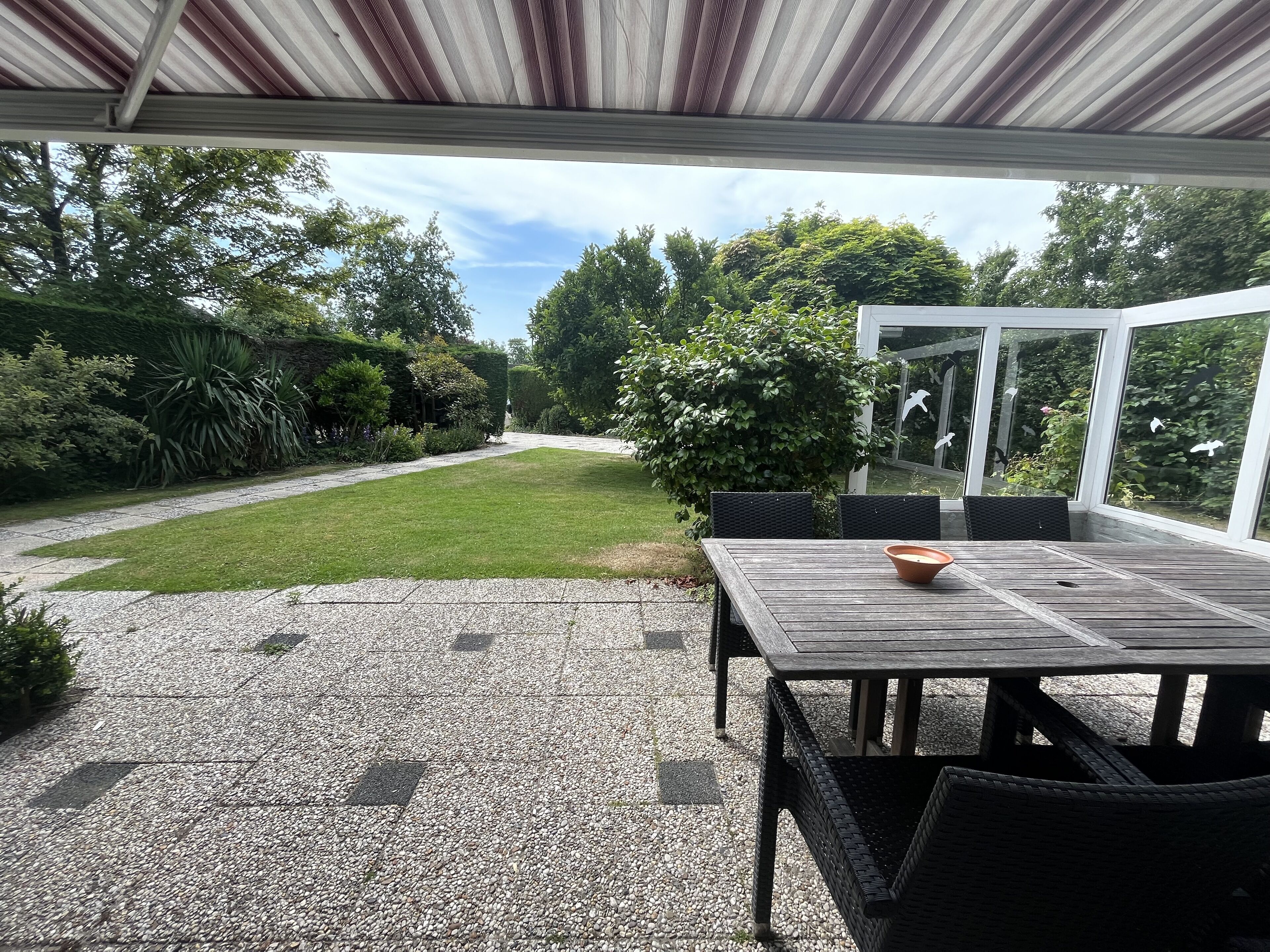 Une terrasse couverte avec une table et des chaises, entourée d’un jardin et offrant une vue sur les arbres.