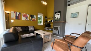 Una sala de estar moderna con un sofá de cuero negro, una mesa de centro de madera y un sillón de cuero marrón. Hay una chimenea con una obra de arte enmarcada arriba, y un televisor en un estante.