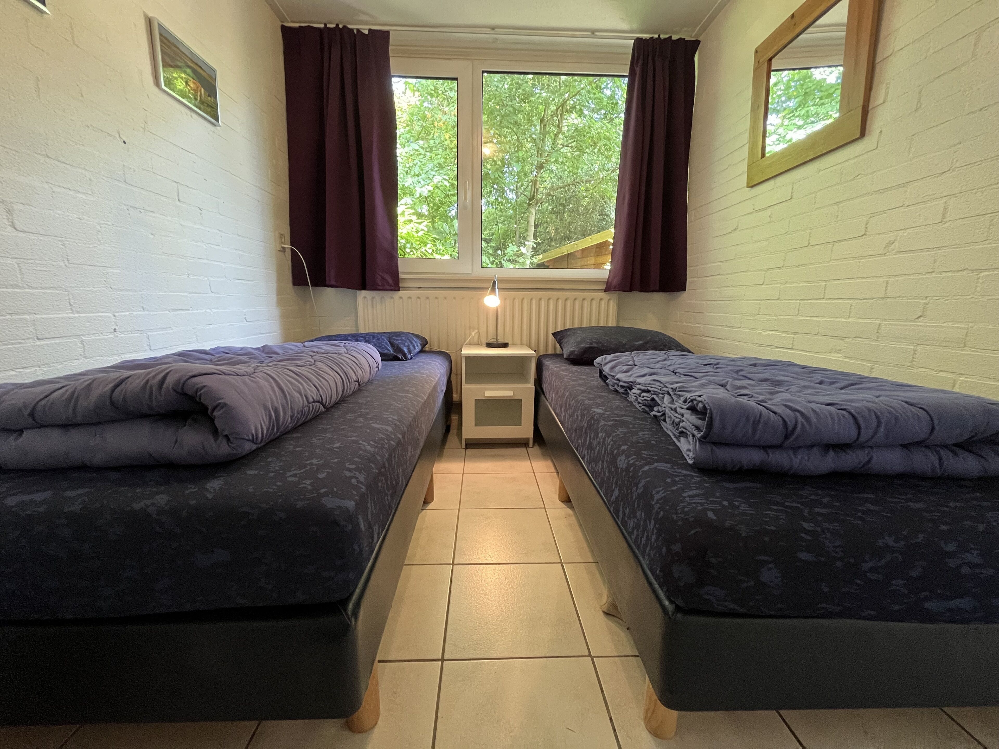 A small, cozy room with two beds, a window with curtains, a radiator, and a small table.