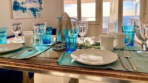 Taula de dinar posta amb vetrería blava, plats blancs i coberts, amb vista al mar a través de grans finestres.