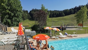 A swimming pool area with lounge chairs, umbrellas, and a playground in the background. - Hostel Quartier Libre (Saint-Jean-en-Royans)