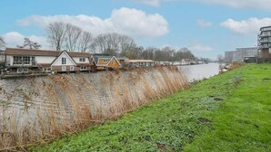 A riverside view with houses along the water, a grassy embankment, and a clear sky.