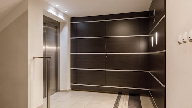 Ein moderner Aufzug mit Metalloptik, neben einer Wand mit horizontalen Holzpaneelen und einem Fliesenboden.