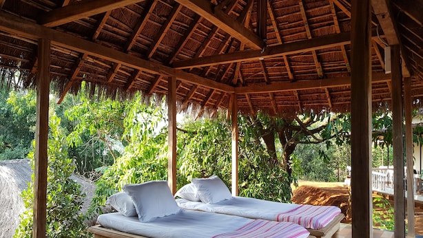 A covered outdoor seating area with two wooden beds, a tiled floor, and a thatched roof.