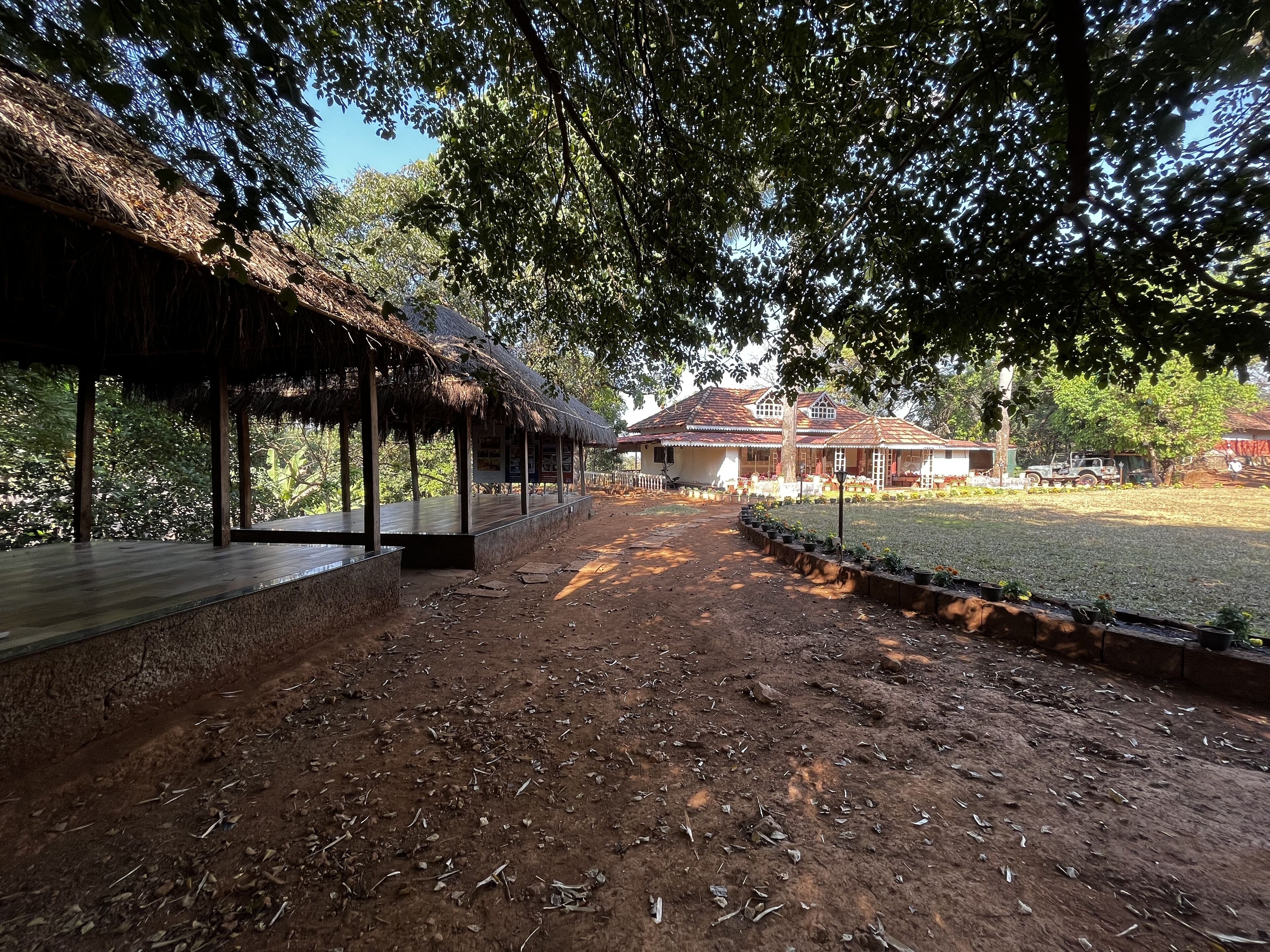 A rustic thatched-roof pavilion with a wooden deck, surrounded by a dirt path and lush greenery.