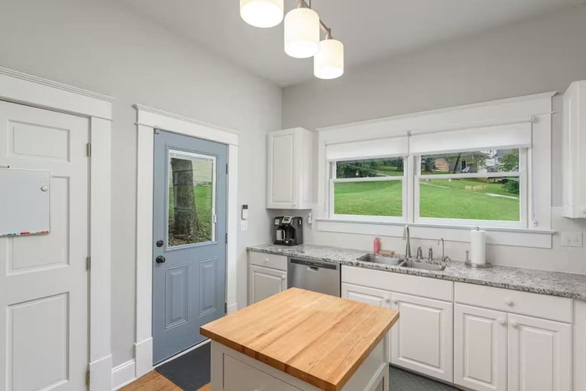 A kitchen with white cabinets, a wooden countertop, a coffee maker, and a window with a view of a green lawn.