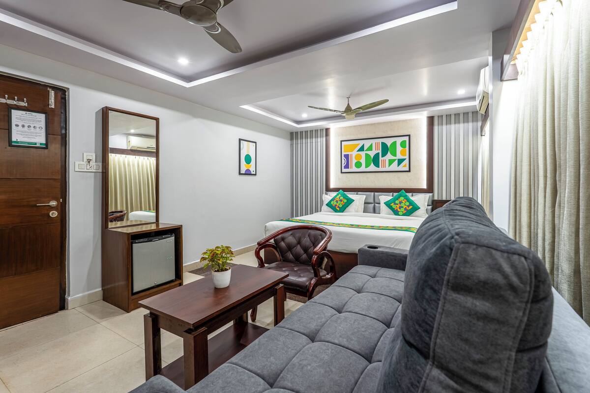 Una habitación de hotel moderna con sofá, sillón, mesita, ventilador de techo y una cama con almohadones decorativos.