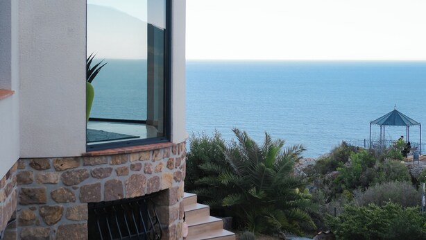 Ein modernes Haus mit Natursteinfassade, einem großen Fenster mit Meerblick und einer Treppe zu einer Terrasse.