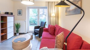 A living room with a red sofa, a black floor lamp, a wooden cabinet, and a window with curtains. - Die Ferienwohnung Seute Deern in St. Peter-dorf für bis zu 2 Personen (St. Peter-Dorf)