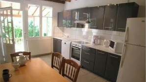 A kitchen with dark cabinets, a white refrigerator, a microwave, and a stove. There is a dining table with chairs and a window with a view of greenery outside. - Les Roseries Boutique Gite with Garden and Pool (CLUSSAIS LA POMMERAIE)