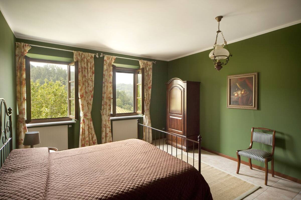 Ein Schlafzimmer mit einem Bett, einem Sessel, einer Heizung, einem Fenster mit Vorhängen, einem Holzschrank und einem Bild an der Wand.