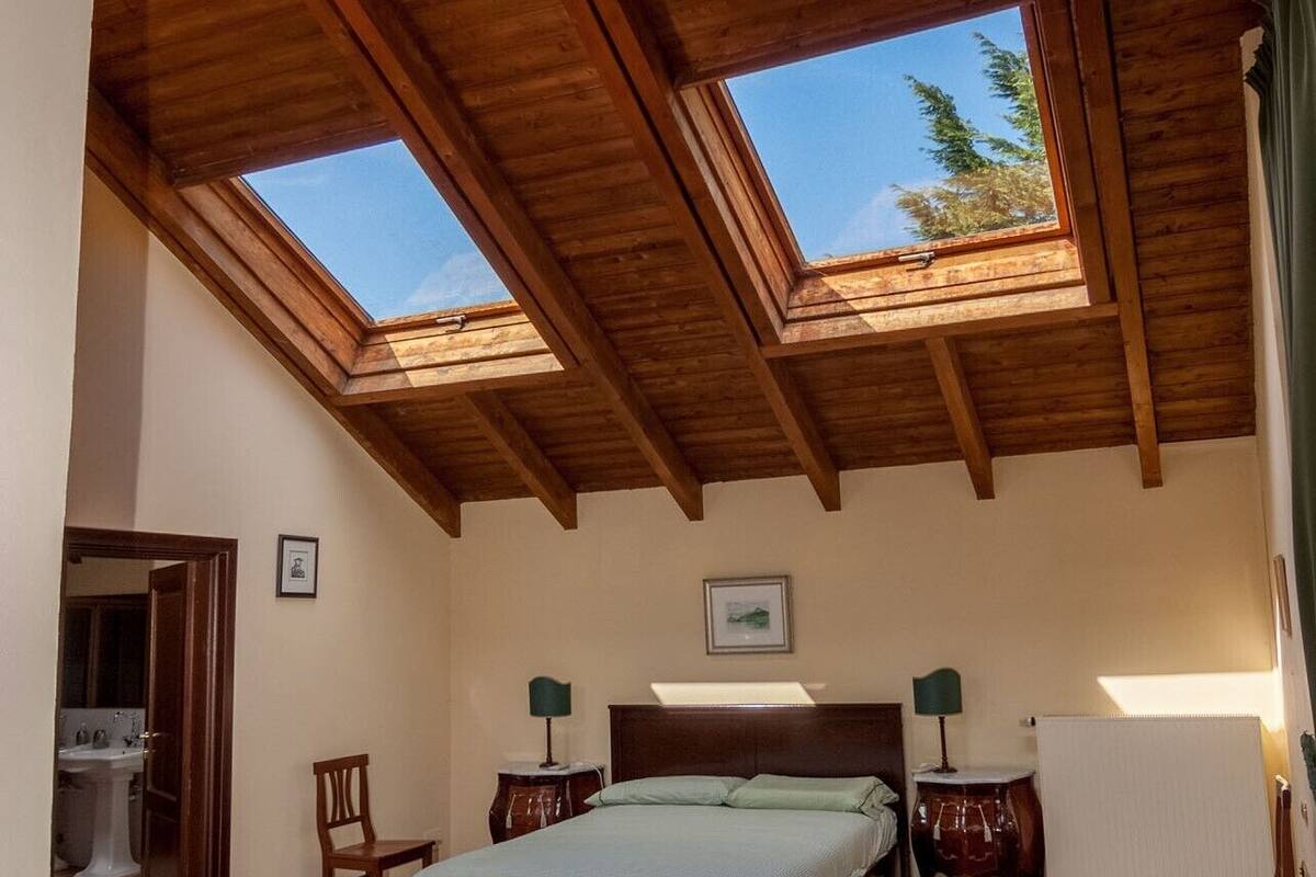 Ein Schlafzimmer mit Holzbalkendecke, Oberlichtern, einem Bett, zwei Nachttischen, einem Sessel und einer Heizung.
