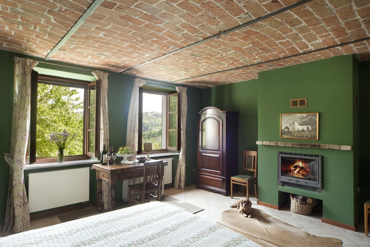 Ein Zimmer mit Ziegeldecke, grünen Wänden, einem Kamin, einer großen Holzkassette und einem mit Vorhängen versehenen Fenster.