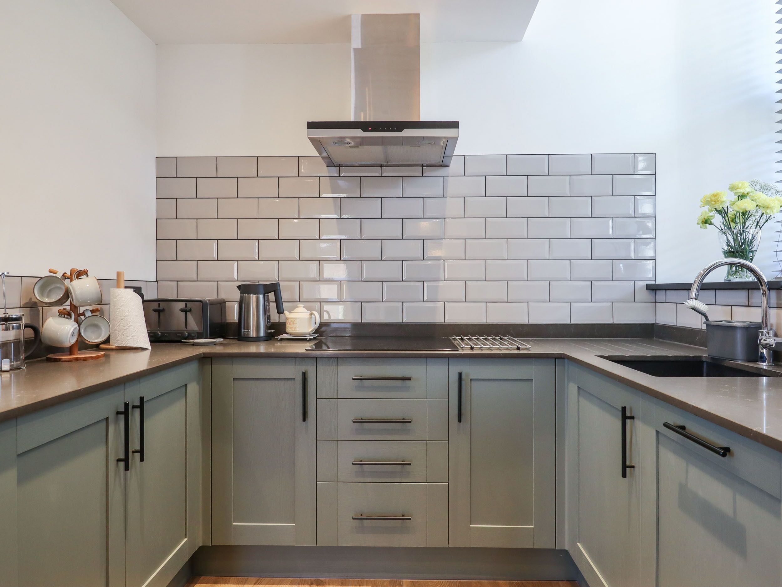 A modern kitchen with grey cabinets, a stainless steel range hood, and a tiled backsplash.