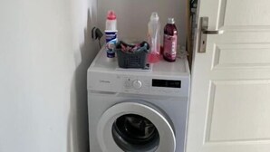 A compact, white washing machine in a narrow laundry room with a door on the right and a window on the left.
