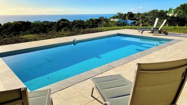 A rectangular pool with clear blue water, surrounded by a tiled deck. There are lounge chairs and a small table on the deck. The background features a view of the ocean and a hilly landscape with greenery.
