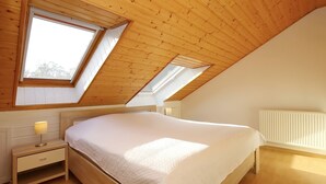 Una camera da letto con soffitto spiovente, lucernari, un letto con copripiumino bianco, un comodino con una lampada e un termosifone.