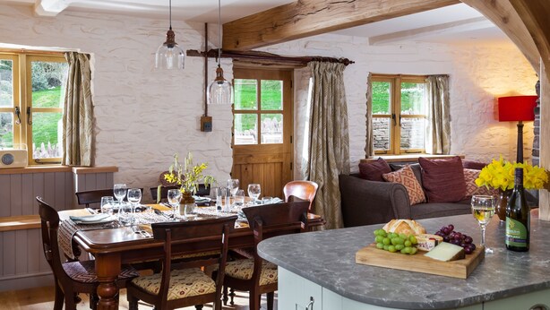 A dining area with a wooden table set for four, a sofa in the background, and a kitchen island with a fruit platter.