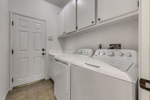 A laundry room with a front-loading washer and dryer, white cabinets above, and a door with a handle. - Stardust: Pool, Spa, FirePit, BBQ, EV Charger! (Indio)