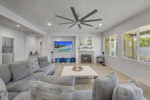 A modern living room with a grey sofa, a fireplace, a flat-screen TV, and a ceiling fan. - Stardust: Pool, Spa, FirePit, BBQ, EV Charger! (Indio)