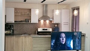 Une cuisine moderne avec des meubles blancs, un îlot central et des luminaires suspendus. Un téléviseur à écran plat est fixé sur un meuble blanc dans l’espace salon.