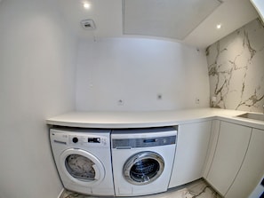 Uma lavanderia com duas máquinas de lavar de abertura frontal, uma bancada de mármore e um balcão branco.