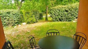 An outdoor patio with a round table and four chairs, surrounded by a garden with various plants and trees. - Welcoming and comfortable T2 house in Fayence (Fayence)