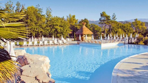 Une piscine bordée de chaises longues et abritée par une structure à toit de chaume, le tout entouré d’arbres et offrant une vue sur des collines lointaines.