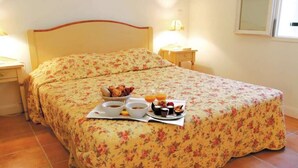Un lit bien fait, recouvert d’une couvre-lit à motifs floraux, une table de chevet avec une lampe et un plateau contenant divers éléments pour le petit-déjeuner, notamment du pain, des fruits et des boissons.