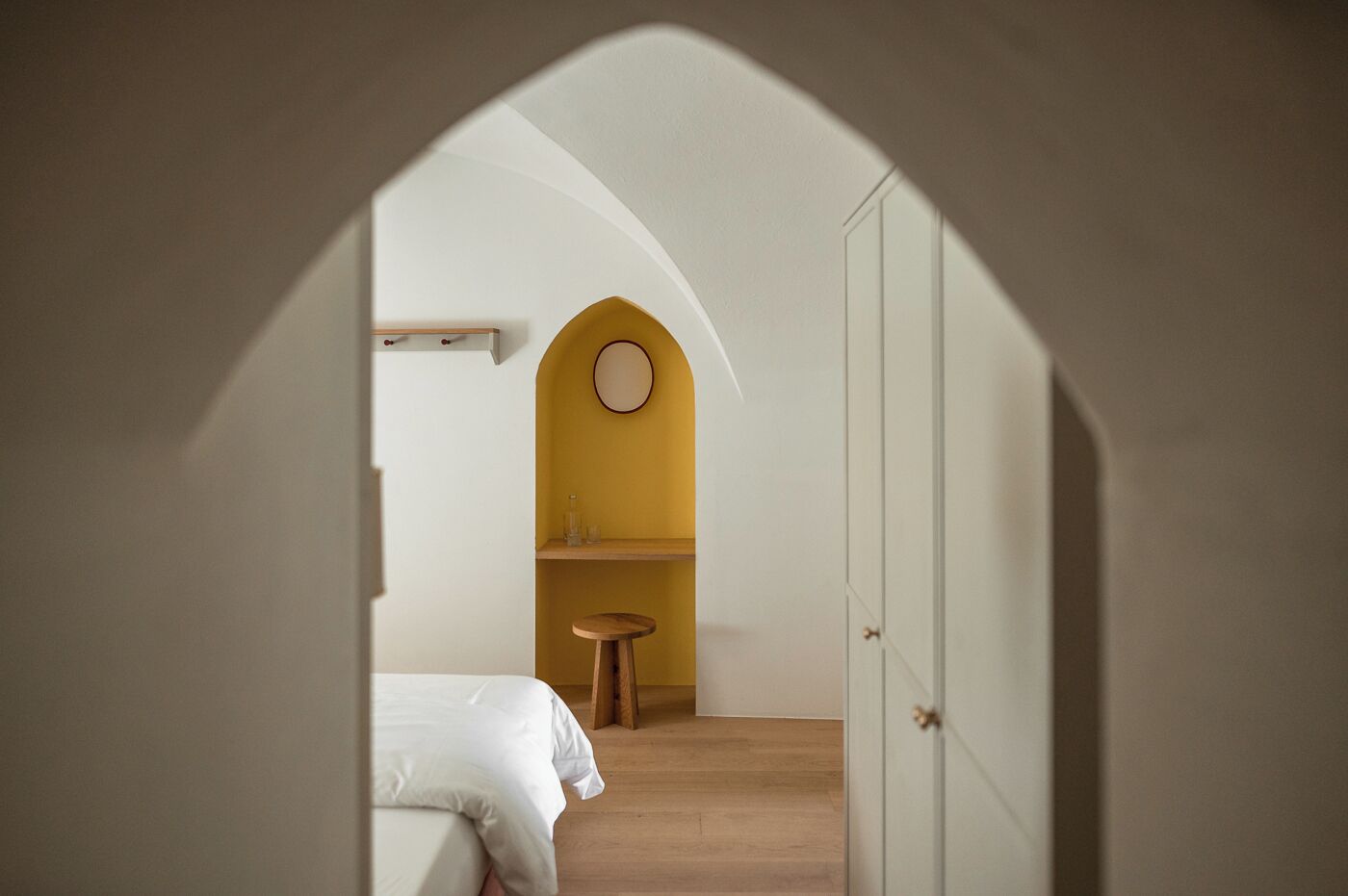 Une chambre à coucher avec un lit, un petit tabouret, un miroir rond et une armoire blanche.