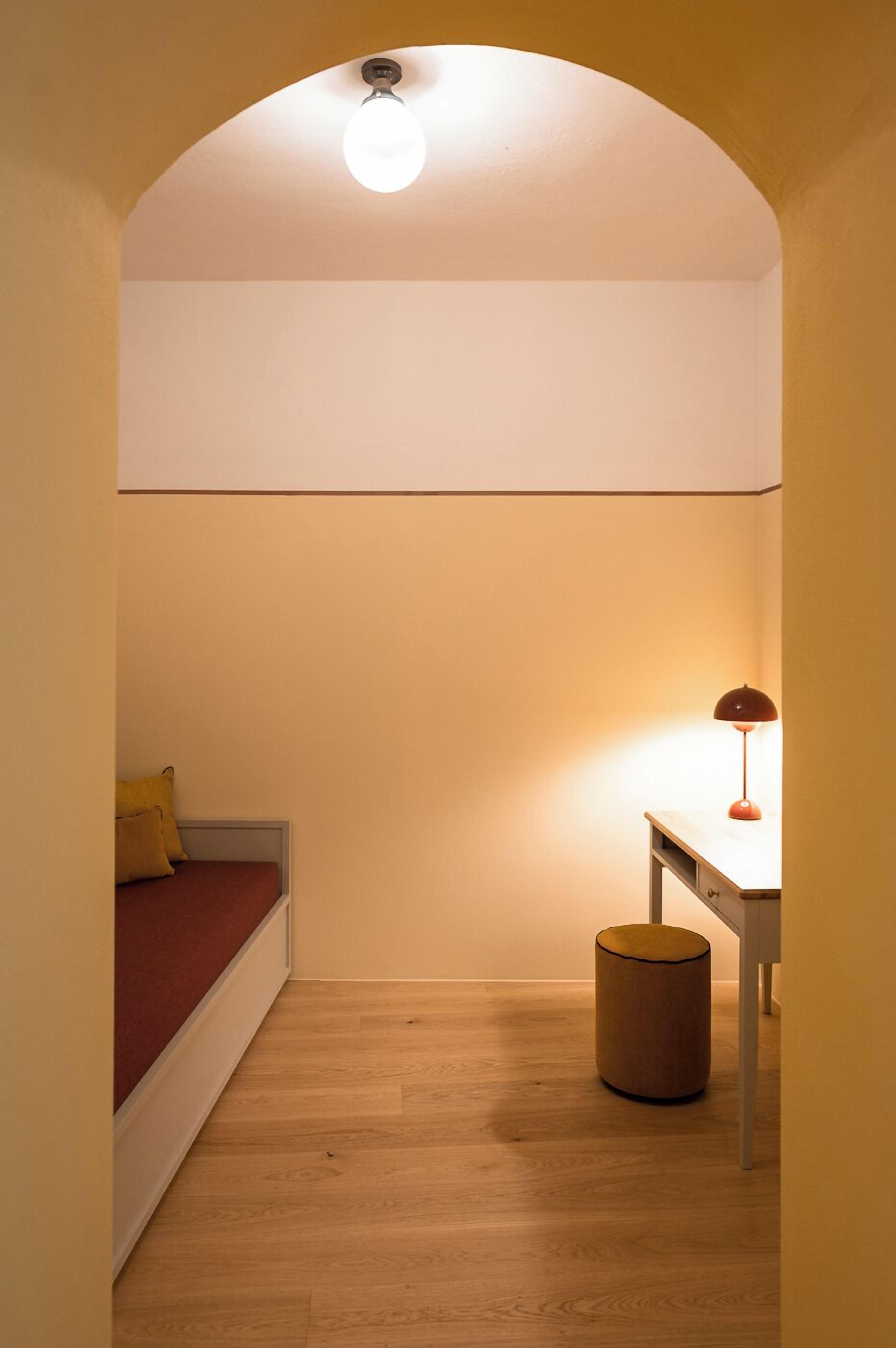 A narrow room with a wooden floor, a bed with red bedding, a desk with a lamp, and a small round stool.