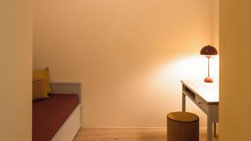A narrow room with a wooden floor, a bed with red bedding, a desk with a lamp, and a small round stool.