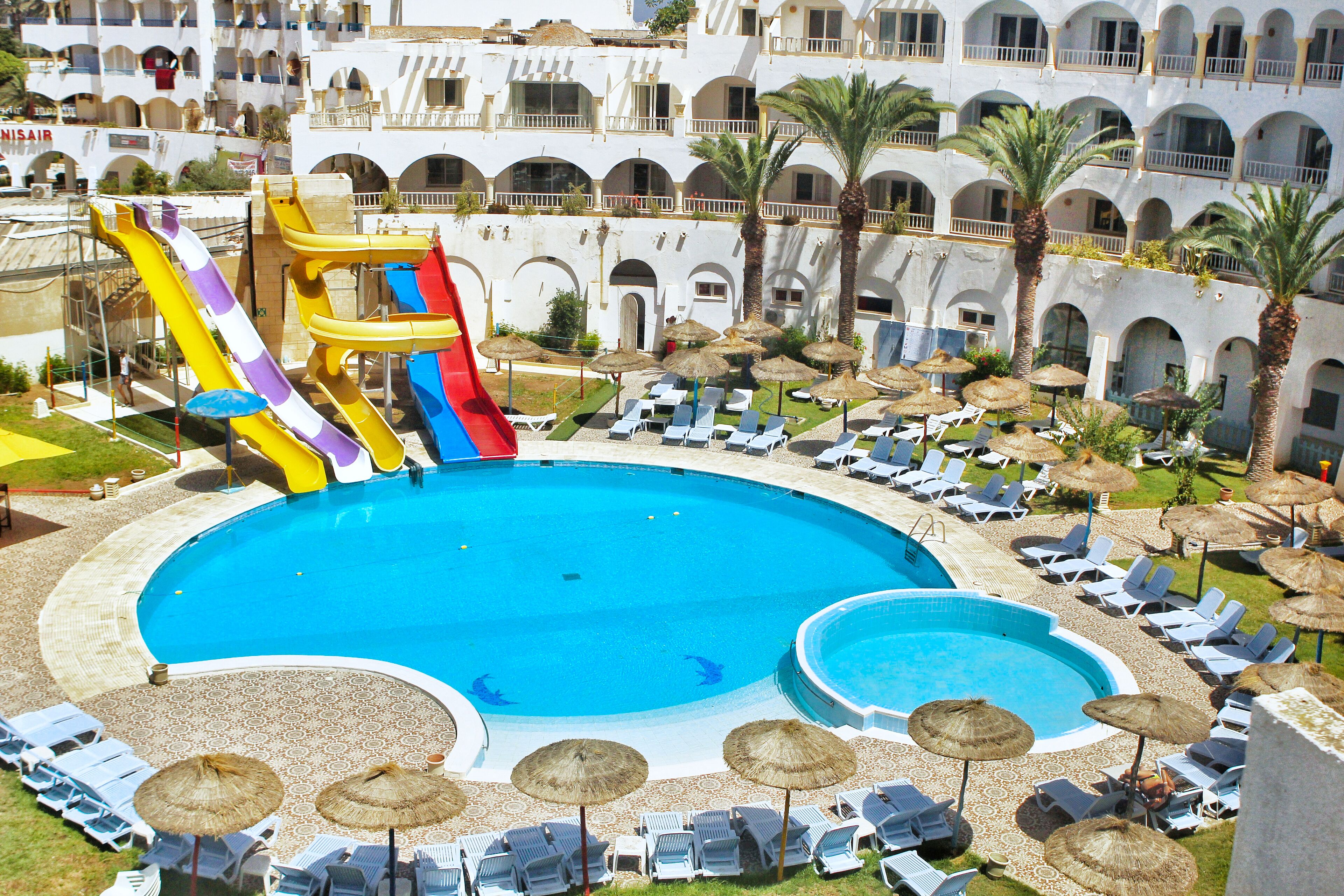 A pool area with slides, lounge chairs, umbrellas, and a building with balconies.